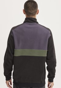 Suéter de forro polar con cuello alto; cuerpo negro, sección superior morada y franja horizontal verde. Presenta mangas largas y un corte holgado.