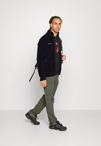 Black fleece jacket with zip, green cargo pants, and black hiking shoes. Features a red graphic shirt, matching tones, and backpack straps.