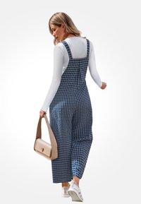 Blauw-witte geruite overall jurk, losjes passend met schouderbanden, gecombineerd met een witte blouse met lange mouwen en een beige handtas. Witte sneakers.