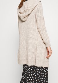 Beżowy dziergany kardigan z kapturem, o luźnym kroju, z prążkowanymi mankietami i dwiema bocznymi kieszeniami. Całość z teksturowanym wzorem.