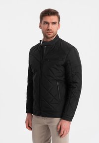 Sort quiltet jakke med opretstående krave, med lynlås fortil, sidelommer og en skrå brystlomme.