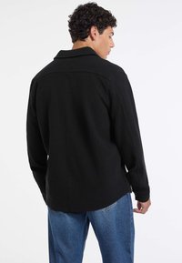 Uomo con capelli ricci, che indossa una maglietta a maniche lunghe nera e jeans blu, di spalle contro uno sfondo bianco semplice.