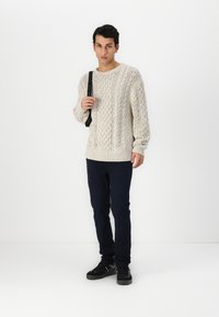 Πλεκτός πουκαμίσα σε ανοιχτό μπεζ με υφή, συνδυασμένος με σκούρους τζιν και μαύρα αθλητικά παπούτσια. Το μοντέλο κρατάει μια τσάντα στον έναν ώμο.