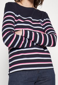 Haut à manches longues bleu marine avec des rayures horizontales rose, bleu clair et blanche. Il présente un col rond et une coupe ajustée.