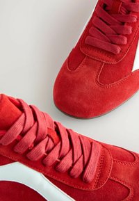 Red suede sneakers with matching laces and white side accents, featuring a rounded toe and textured upper for a sporty design.