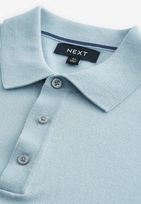 Lyseblå polo shirt lavet af tekstureret stof. Den har en krave, en tre grå knapper lukning og en kontrasterende stribe indeni kraven.