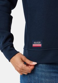 Marineblauwe sweatshirt met ribgebreide manchetten en zoom; heeft een rechthoekig label met "SEASIDE" en een rood-wit-blauw streepdesign.