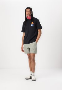 Sort kortærmet T-shirt med farverig grafik på venstre bryst, parret med lysegrønne shorts. Modellen bærer et rødt bandana og sneakers.
