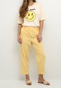 T-shirt crème avec un motif de smiley jaune, associé à un pantalon léger jaune taille haute et des sandales plates beiges.