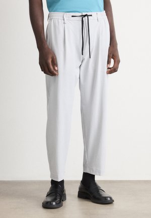 Man in een lichtgrijze verstelbare broek, met zwarte sokken en zwarte leren instap schoenen, staande op een lichte vloer tegen een effen muur.