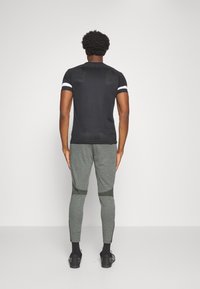 Camisa negra ajustada de manga corta con rayas blancas en las mangas, combinada con pantalones grises claros ajustados que presentan detalles en verde oscuro.