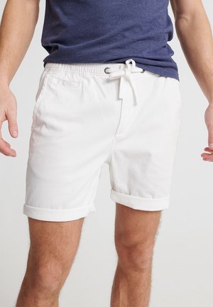 Homme portant une chemise bleu marine et un short blanc à cordon avec ourlets retroussés, debout devant un fond uni.
