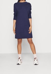 Robe en coton bleu marine avec une coupe décontractée, des manches longues et des poignets côtelés. Portée avec des baskets blanches et des chaussettes roses sur un fond uni.