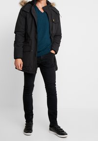 Mustallinen takki, jossa on turkisvuorattu huppu, kerroksittain teal-värisen neuleen päällä. Yhdistetty tiukkoihin mustiin farkkuihin ja mustiin lenkkareihin, joissa on hihnakiinnitykset.