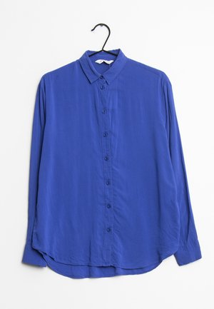 Chemise bleu roi à manches longues avec boutons et col, suspendue sur un cintre noir sur fond blanc.
