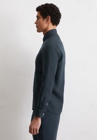 Donkergroene blouse met lange mouwen in een linnenmix, voorzien van een knop-onder hals en kleine knopen bij de manchetten, zijwaarts weergegeven.