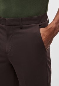 Calças de tecido castanho com textura suave, bolsos laterais e corte justo. A cintura inclui passadores para cinto, permitindo ajustar o tamanho.