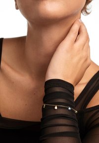 Bracciale in catena d'oro con pendenti di pietre preziose colorate. Indossato su una sopra a maniche lunghe trasparenti nere, con polsini aderenti.
