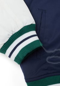 Cuff di una bomber jacket blu e bianca con maglina a coste verde e accenti a righe, realizzata in tessuto liscio e lucido.