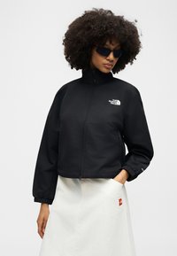 Žena s kudrnatými vlasy, která má na sobě černé sluneční brýle, černou bundu na zip s logem „The North Face“ a bílou sukni, ruka v kapse.