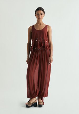 Mujer con un top sin mangas de color rojo óxido con volantes y adornos, combinado con pantalones sueltos y transparentes a juego y sandalias de plataforma.