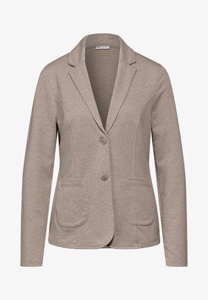 Mujer con blazer beige sobre camisa blanca y vaqueros azules, con una mano en el bolsillo contra un fondo gris liso.