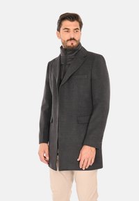 Uomo che indossa un cappotto grigio scuro su misura sopra una giacca nera con cerniera e pantaloni beige, in piedi contro uno sfondo chiaro uniforme, che guarda di lato.