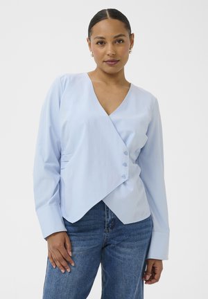 Kobieta związaną z tyłu fryzurą, ubrana w jasnoniebieską bluzkę o kopertowym kroju z trzema guzikami, połączoną z niebieskimi dżinsami.