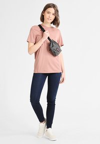 Grå tekstureret bæltetaske båret over en lyserød t-shirt, parret med mørkeblå jeans og hvide sneakers, stående foran en ensfarvet baggrund.