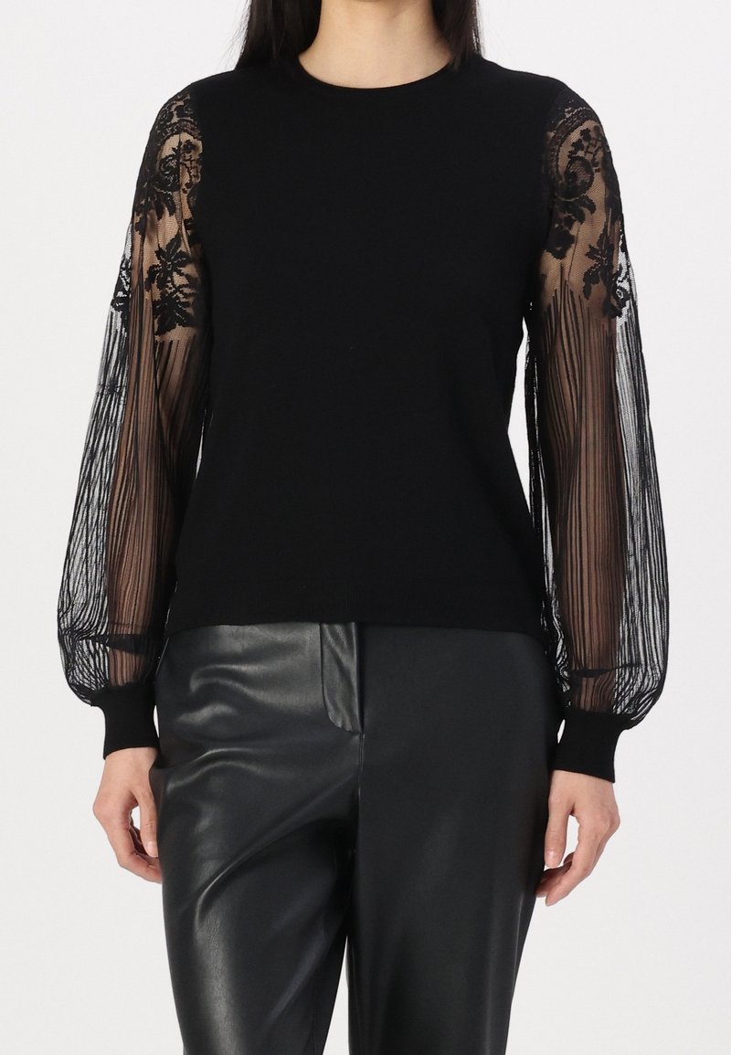 Haut noir avec manches bouffantes en dentelle florale transparente, associé à un pantalon en cuir noir porté par une personne debout.