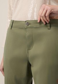 Olivgröna byxor med en slät textur, som har dragkedja, knäppning och bältesöglor. Ytterligare stickningsdetalj längs fickorna.