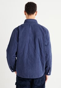 Chemise matelassée bleue avec un motif de coutures courbes, poignets boutonnés et col. Le tissu semble doux et texturé, idéal pour une tenue décontractée.