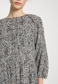 Mekko, jossa on musta-valkoinen kuviointi, pyöreä pääntie, rypistetyt yksityiskohdat ja kyynärpäihinsulkeutuvat hihat, sekä virtaava, laskeutuva muotoilu.