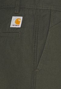 Olijfgroene stoffen broek met zichtbare stiksels, zijzakken en een wit label met een oranje logo. Duurzaam, arbeidskleding ontwerp.