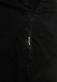Tessuto nero con una sottile texture, caratterizzato da un'etichetta con cucitura laterale con il testo "CHASIN'" in bianco. Il design è semplice e utilitario.