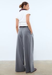 Femme avec les cheveux en chignon portant une chemise blanche à manches courtes et un pantalon large gris, les mains dans les poches, debout sur un sol en béton.