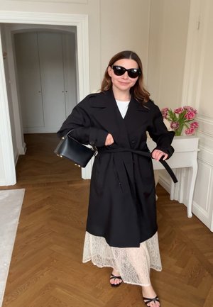 Kvinde iført sort trenchcoat, solbriller, bærende på sort håndtaske, binder bælte i et rum med trægulv og hvide vægge, ved siden af et hvidt bord med pink blomster.