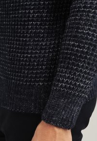 Ciemnoniebieski sweter z dzianiny o teksturowanym wzorze, noszony na ramieniu osoby na tle w neutralnych kolorach.