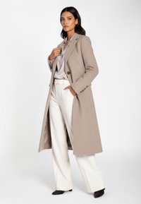 Beige lång kappa i texturerat tyg med knäppning med en knapp och sidfickor, kombinerad med en ljus blus och vita vida byxor.