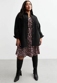 Schwarze oversized Jacke mit Knopfverschluss über einem pinken gemusterten Kleid. Knielange schwarze Stiefel vervollständigen das Outfit. Glatte Textur überall.