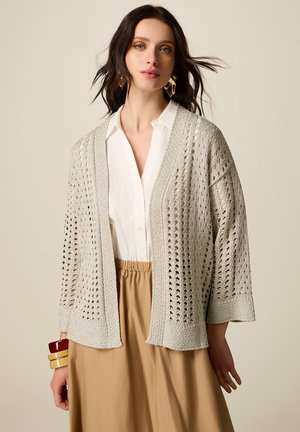 Vrouw draagt een beige opengebreid vest over een witte blouse en een beige rok, gecombineerd met gouden oorbellen en kleurrijke armbanden aan de linkerpols.