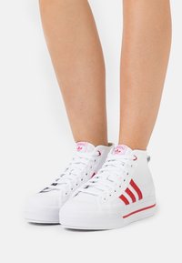White high-top sneakers made of smooth leather with red stripes, textured rubber outsole, and pink logo tag at the tongue.