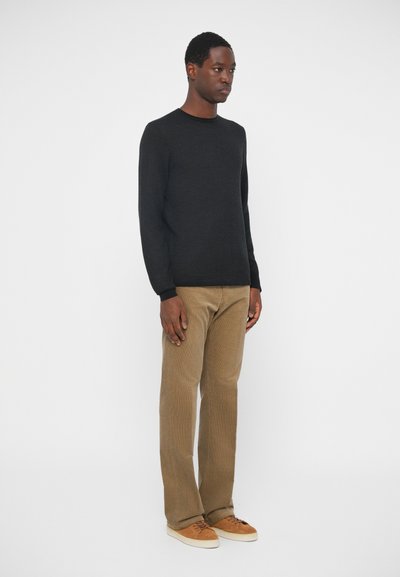 Black crew-neck knit sweater, beige corduroy trousers, and beige sneakers. Model poses against a plain white background.