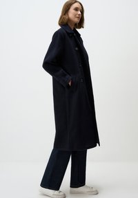 Marineblauwe jas van een gestructureerde stof, met een kraag, knoopsluitingen en zijzakken, gecombineerd met uitlopende denim broek en witte sneakers.