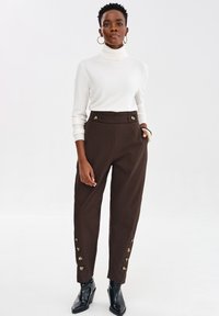 Kobieta ubrana w biały sweter z golfem, brązowe spodnie z wysokim stanem z złotymi guzikami po bokach oraz czarne, szpiczaste botki za kostkę, stojąca na tle białym.