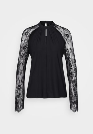 Blouse noire avec de longues manches en dentelle, col montant et détail en forme de goutte d'eau à l'avant. Le tissu du corps lisse contraste avec la texture en dentelle complexe.