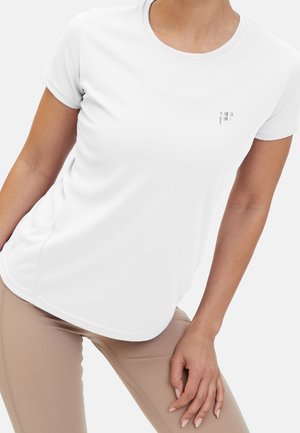 Vrouw draagt een nauwsluitend wit sportshirt met korte mouwen en beige leggings, poseert met één hand op haar dij tegen een witte achtergrond.