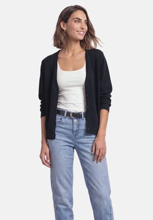 Frau trägt schwarzen, geknöpften Cardigan, weißes Tanktop, hellblaue Jeans und schwarzen Gürtel, lächelt und schaut seitlich vor weißem Hintergrund.