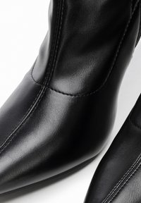 Detailní záběr na černé kožené kotníkové boty s špičatými špičkami a viditelnými švy na bílém pozadí.