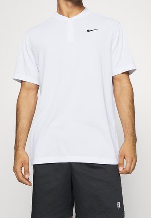 Mężczyzna w białej koszulce Nike z krótkim rękawem i czarnych spodenkach, stojący na tle jednolitego, jasnego tła.